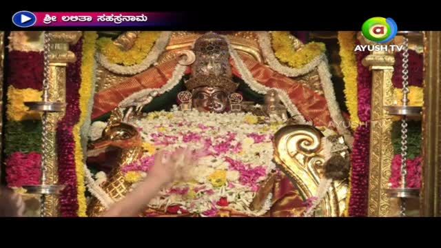 A hand gently showers flower petals over a bedecked deity, the scene broadcast by Ayush TV from India. The camera focuses on the gilded details and the layers of floral offerings.

