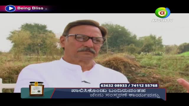 A man in glasses and a white shirt is speaking on Ayush TV, with a jar of honey displayed on the screen. Behind him, a field and some trees are visible.
