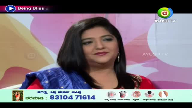 A woman with dark hair and a blue shawl is seated on a set for Ayush TV in India. The show's title, "Being Bliss," is displayed in the corner.
