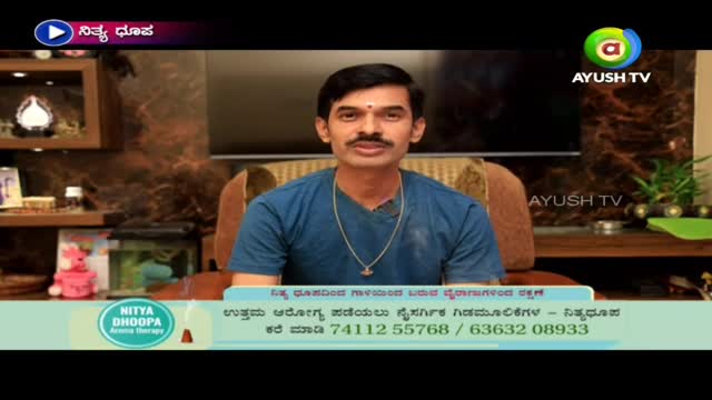 A man with a mustache and a bindi is seated in front of a camera, speaking on Ayush TV. Below him, a graphic advertises "Nitya Dhoopa" and lists a phone number.
