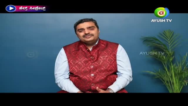 A man in a patterned red vest and white shirt sits facing the camera, likely on an Ayush TV program. The background is a solid blue, with the Ayush TV logo visible.
