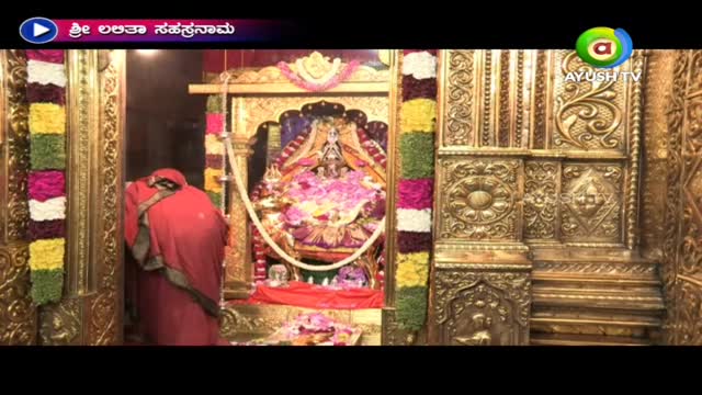 A person in a red sari bows before a deity inside a temple, framed by elaborately carved golden doors.  The scene is being broadcast on Ayush TV, with a title card in the upper left corner.
