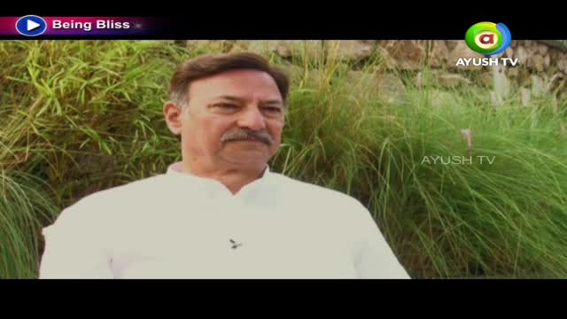 A man in a white shirt sits in front of a wall of tall grass, the Ayush TV logo visible in the corner. He's likely on a show called "Being Bliss" in India.
