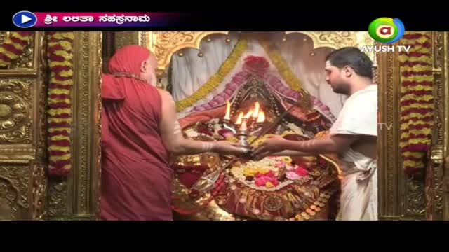 Two men in traditional garb are holding a small, ornate lamp with flames in front of a decorated altar. The scene is being broadcast on Ayush TV, with a title card in the upper left corner.
