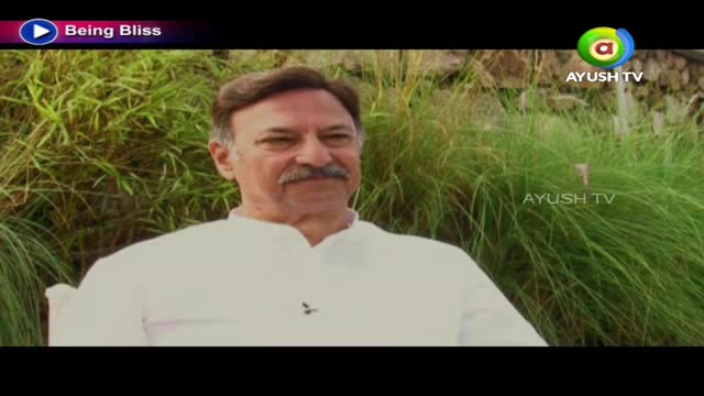 A man with a mustache sits in a white shirt, looking toward the camera. Behind him, tall green grass and a logo for Ayush TV are visible.

