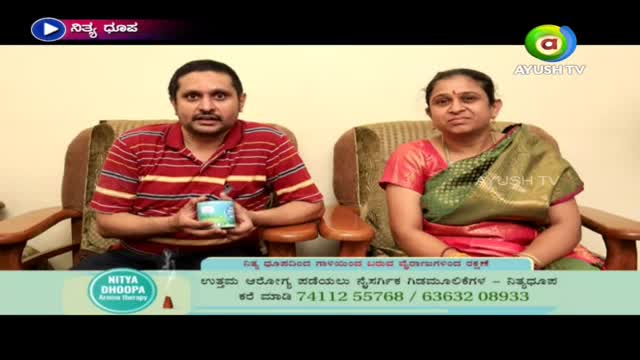 A man in a red and white striped shirt holds a small box, speaking on what appears to be an Ayush TV program. Beside him, a woman in a red sari and green shawl sits, listening.
