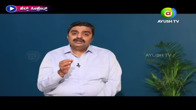 A man in a light blue shirt gestures with his right hand while speaking, likely on the Ayush TV program. The backdrop is a simple blue wall with a small plant visible on the right side of the frame.
