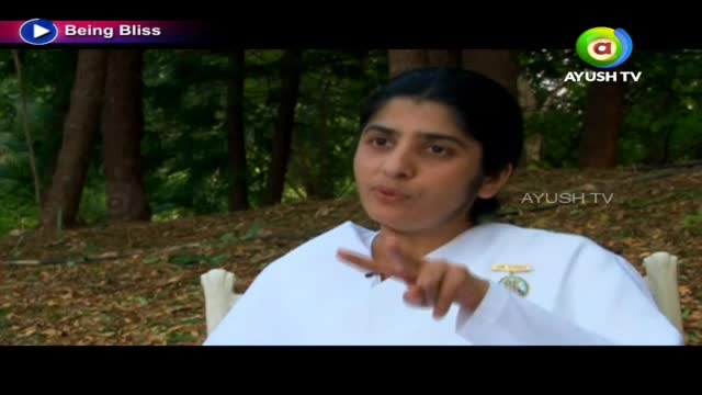 A woman in a white robe gestures with her right hand while speaking. The Ayush TV logo is visible in the corner, and the background is a wooded area.

