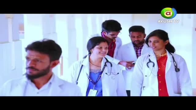 A group of people in white coats and stethoscopes walk down a bright hallway, smiling and talking. The Ayush TV logo sits in the upper right corner.
