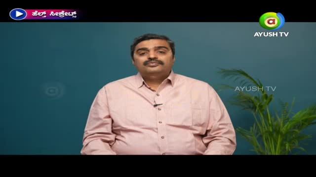 A man in a pink shirt speaks directly to the camera, a small microphone clipped to his collar. The Ayush TV logo is visible in the corner, and a plant sits to the right.
