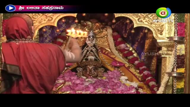 A person in a red robe holds a lit candle towards a deity adorned with flowers and jewels. This is likely a broadcast on Ayush TV from India, as the channel's logo is visible in the corner.
