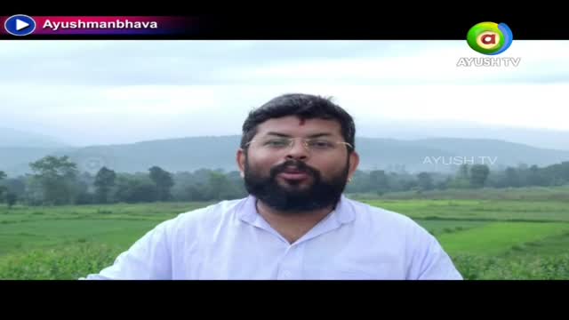 A man with a full beard and glasses is centered, speaking in front of a green field and distant hills. The Ayush TV logo is visible in the corner, and "Ayushmanbhava" is displayed at the top.
