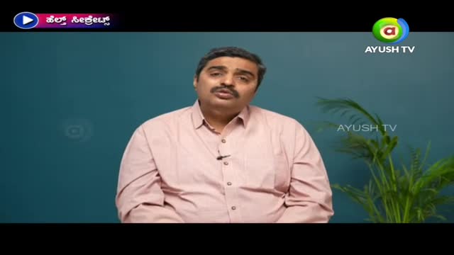 A man in a pink shirt speaks directly to the camera, a small microphone clipped to his collar. The Ayush TV logo is visible in the corner of the screen, and a plant sits to the right.
