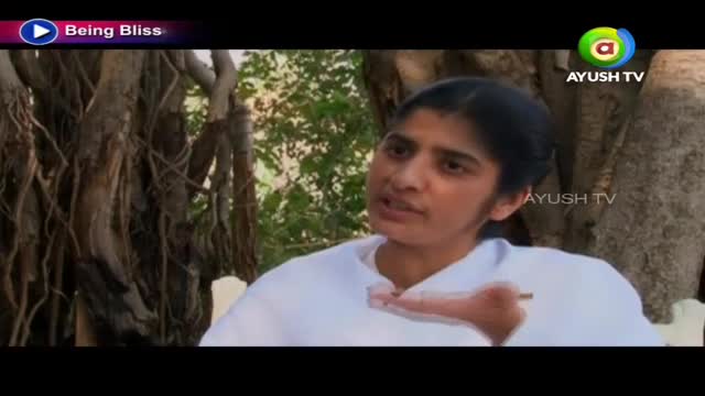 A woman in a white top gestures with her right hand while speaking, likely for Ayush TV. Behind her, the gnarled roots of a large tree are visible.
