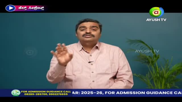 A man in a pink shirt gestures with his right hand, speaking on Ayush TV. Behind him, a simple teal backdrop and a small plant provide a clean setting.
