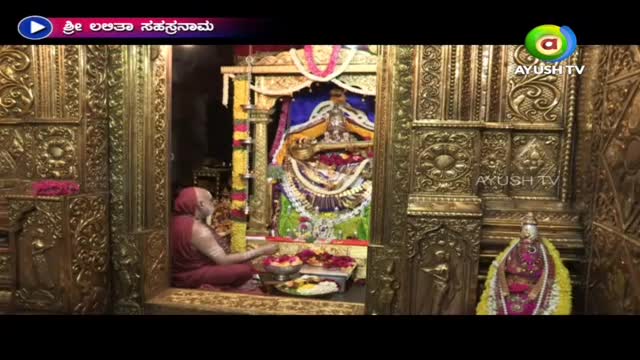 A robed figure sits before an ornate shrine, arranging flowers and other offerings. The scene is broadcast on Ayush TV, with a title card in the upper corner.
