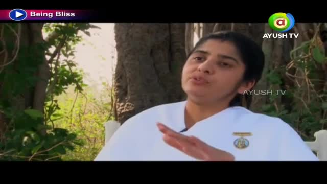 A woman in a white outfit gestures with her right hand, speaking to someone off-camera. The Ayush TV logo is visible in the corner, and the backdrop is a natural setting.
