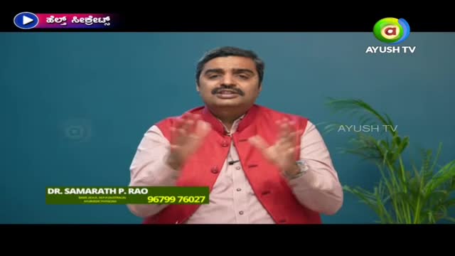 A man in a red vest and white shirt gestures with both hands, speaking on Ayush TV. His name and a phone number are displayed on a lower-third graphic.
