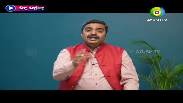 A man in a red vest gestures with his hands while speaking on Ayush TV. The set has a blue background and a small plant to the side.
