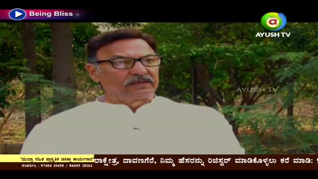 A man in glasses and a white shirt is speaking, the Ayush TV logo visible in the corner. Behind him, green trees create a soft backdrop for the interview.
