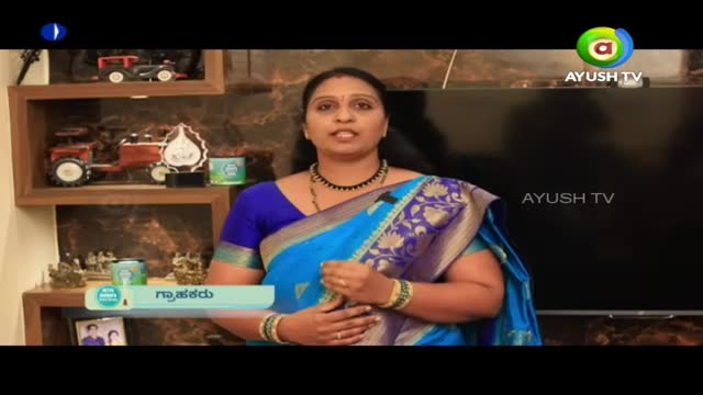 A woman in a blue sari stands in front of a large screen, speaking on Ayush TV. Behind her, a shelf displays model tractors and other decorative items.

