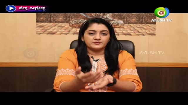 A woman in an orange shirt gestures with her hands as she speaks, likely on a segment for Ayush TV in India. The backdrop is a neutral wall with a framed picture, and the logo for "Health Secrets" is visible in the top left.
