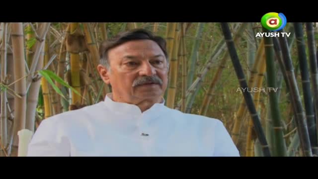 A man in a white shirt is speaking, his gaze directed slightly to the side. Behind him, a screen displays the Ayush TV logo, framed by a bamboo grove.
