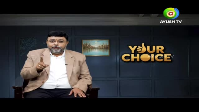 A man in a light-brown blazer sits in a chair, pointing his index finger upwards. The set behind him displays the "Your Choice" logo, with the Ayush TV logo visible in the corner.
