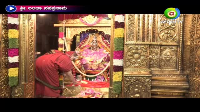 A person in a red robe is carefully arranging flowers at the base of a shrine. The scene is being broadcast on Ayush TV, with the title "Sri Lalitha Sahasranama" displayed at the top.
