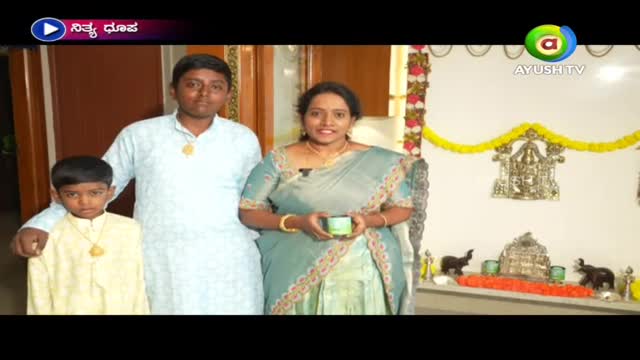 A family of three stands in front of a decorated wall, the woman holding a small green container.  The Ayush TV logo is visible in the upper right corner.
