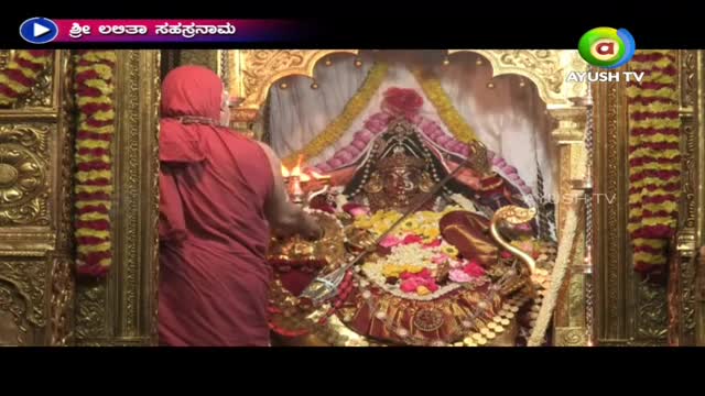 A robed figure in red, seen from the back, tends to an ornate shrine. The scene is broadcast on Ayush TV, likely from a temple in India.
