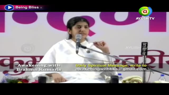 A woman in a white outfit speaks into a microphone, likely on Ayush TV, with the Brahma Kumaris logo visible. The backdrop features large, stylized lettering in a dark color.
