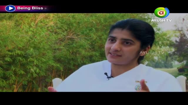 A woman in a white outfit gestures with her hands while speaking. The Ayush TV logo is visible in the corner, and a lush green backdrop fills the space behind her.
