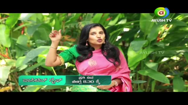 A woman in a pink sari gestures with her right hand, speaking to the camera for Ayush TV. Lush green foliage forms a backdrop behind her.

