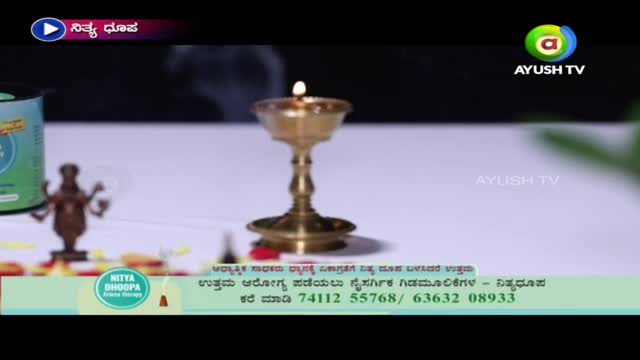 A small oil lamp with a flickering flame sits centered on a white surface. The Ayush TV logo is visible in the corner of the screen, and the words "Nitya Dhoopa" are displayed above the lamp.
