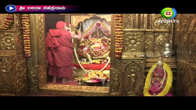 A robed figure in red is reaching into a golden shrine, possibly arranging flowers.  Ayush TV's logo is visible, suggesting a broadcast from India.
