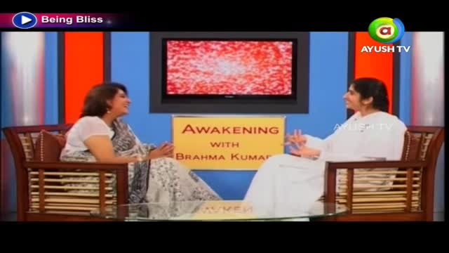 Two women are seated in wooden chairs, engaged in a conversation on a set for Ayush TV. The show, "Awakening with Brahma Kumar," is underway, with a static screen displaying behind them.
