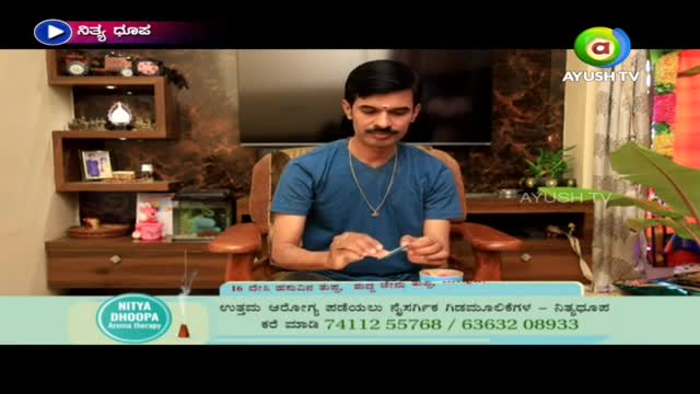 A man in a blue shirt sits in a chair, carefully holding a small piece of paper. He appears to be working with incense, as the Ayush TV logo is visible.
