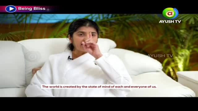 A woman in a white robe sits on a white couch, gesturing with her index finger. The Ayush TV logo is visible in the top right corner.
