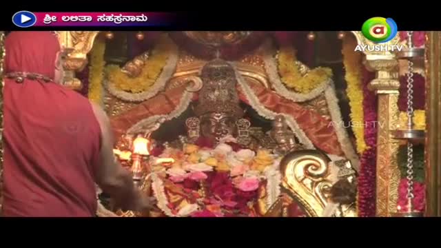 A robed figure, seen from the back, is attending to a shrine adorned with flowers and gold. The broadcast logo for Ayush TV is visible in the corner of the screen.
