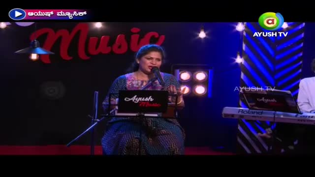 A woman in a patterned sari sings into a microphone, her hands gesturing as she performs. The stage is set for a music show, with the Ayush TV logo visible on the screen and the keyboard.
A woman in a patterned sari sings into a microphone, her hands gesturing as she performs. The stage is set for a music show, with the Ayush TV logo visible on the screen and the keyboard.