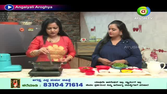A woman in a red top stirs something in a pan on a small burner, while another woman stands nearby. The scene appears to be part of a cooking show on Ayush TV, with ingredients and a water pitcher arranged on the counter.
A woman in a red top stirs something in a pan on a small burner, while another woman stands nearby. The scene appears to be part of a cooking show on Ayush TV, with ingredients and a water pitcher arranged on the counter.