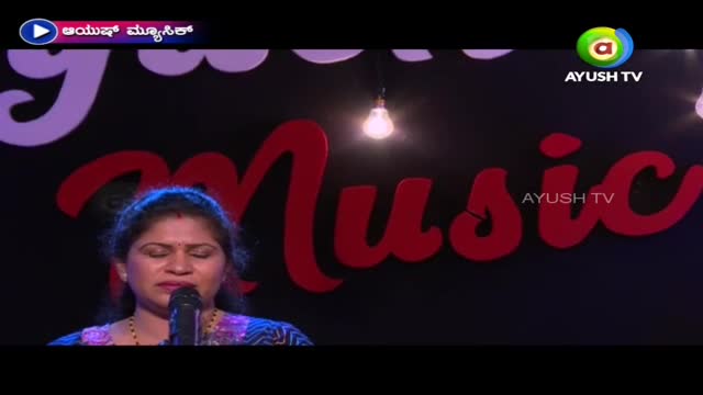 On the Ayush TV stage, a woman sings into a microphone, her eyes closed. Behind her, the word "Music" is spelled out in large, red letters against a dark background.
On the Ayush TV stage, a woman sings into a microphone, her eyes closed. Behind her, the word "Music" is spelled out in large, red letters against a dark background.