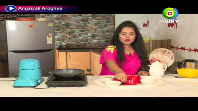 A woman in a bright pink top reaches for a small red bowl on a tray, likely preparing a dish for the Ayush TV show. A pan sits on a small burner, and a blender is next to a refrigerator.
A woman in a bright pink top reaches for a small red bowl on a tray, likely preparing a dish for the Ayush TV show. A pan sits on a small burner, and a blender is next to a refrigerator.
