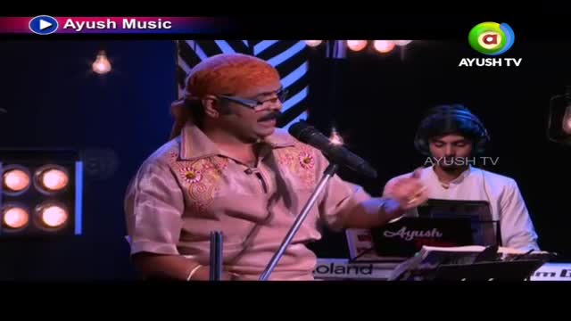 The singer, wearing a turban and glasses, gestures with his right hand as he sings into a microphone. A keyboard player in the background sits with headphones on, ready to play.
