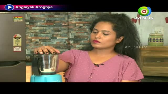 A woman in a pink top is focused on a blender, likely preparing something for the Ayush TV program, "Angaiyali Aroghya." She is holding the lid down, the contents inside hidden from view.
