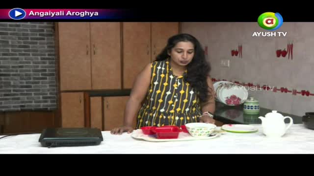 The woman in the patterned dress stands behind a table set with dishes and small red trays. A black hot plate sits to her left, and the Ayush TV logo is visible in the corner.
