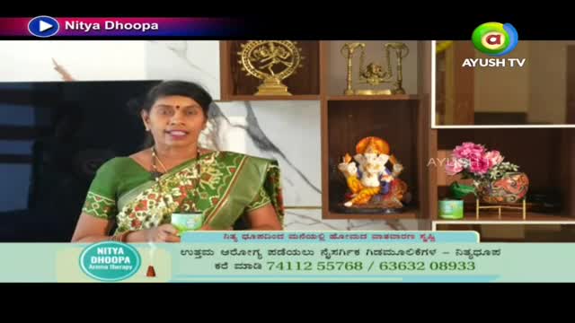 A woman in a green sari is holding a small cup, speaking on a television program called "Nitya Dhoopa" on Ayush TV. Behind her, a shelf displays religious statues and decorative items.
