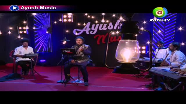 On a stage lit by fairy lights, a musician sings into a microphone while playing a keyboard. Behind him, a band accompanies him, the scene likely part of an Ayush TV music program.

