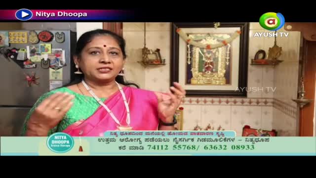A woman in a pink sari gestures with her hands as she speaks, likely on a segment for Ayush TV. Behind her, a framed image of a deity hangs on the wall.
